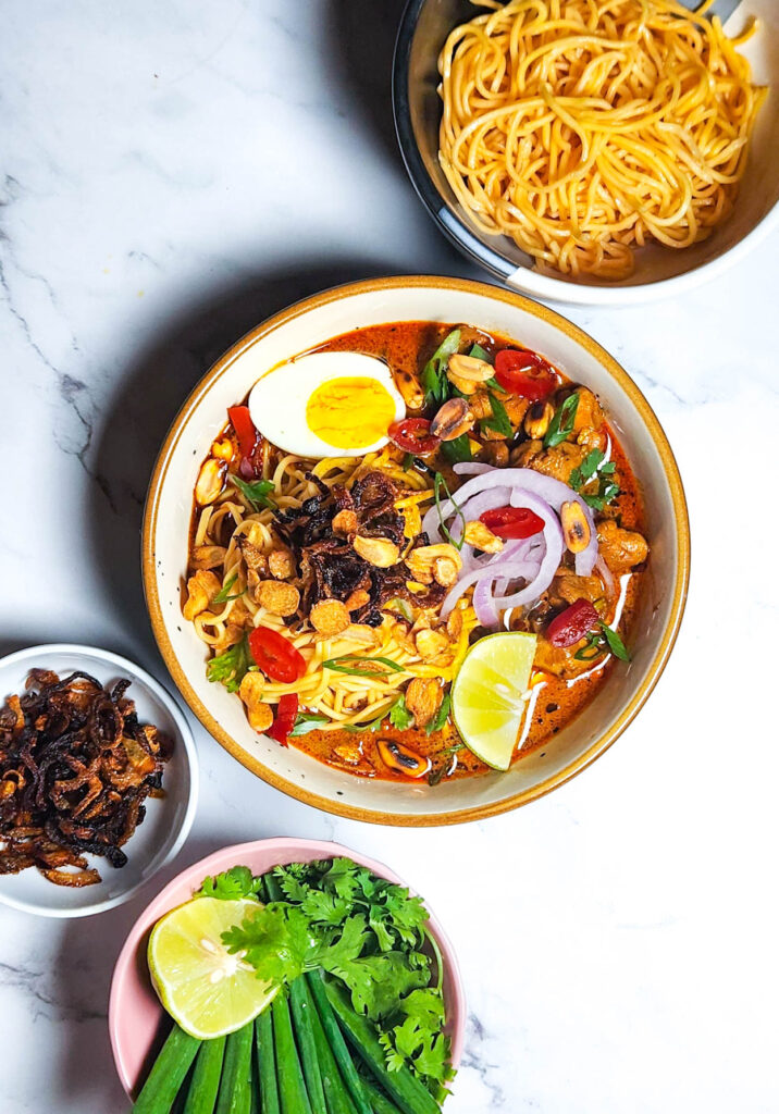 khow suey _ Burmese curry coconut noodle soup in a bowl with toppings lime, boiled eggs, onions, chillies, garlic