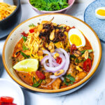 khow suey _ Burmese curry coconut noodle soup in a bowl with toppings lime, boiled eggs, onions, chillies, garlic