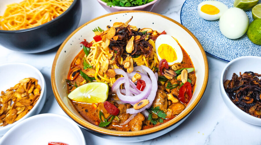 khow suey _ Burmese curry coconut noodle soup in a bowl with toppings lime, boiled eggs, onions, chillies, garlic