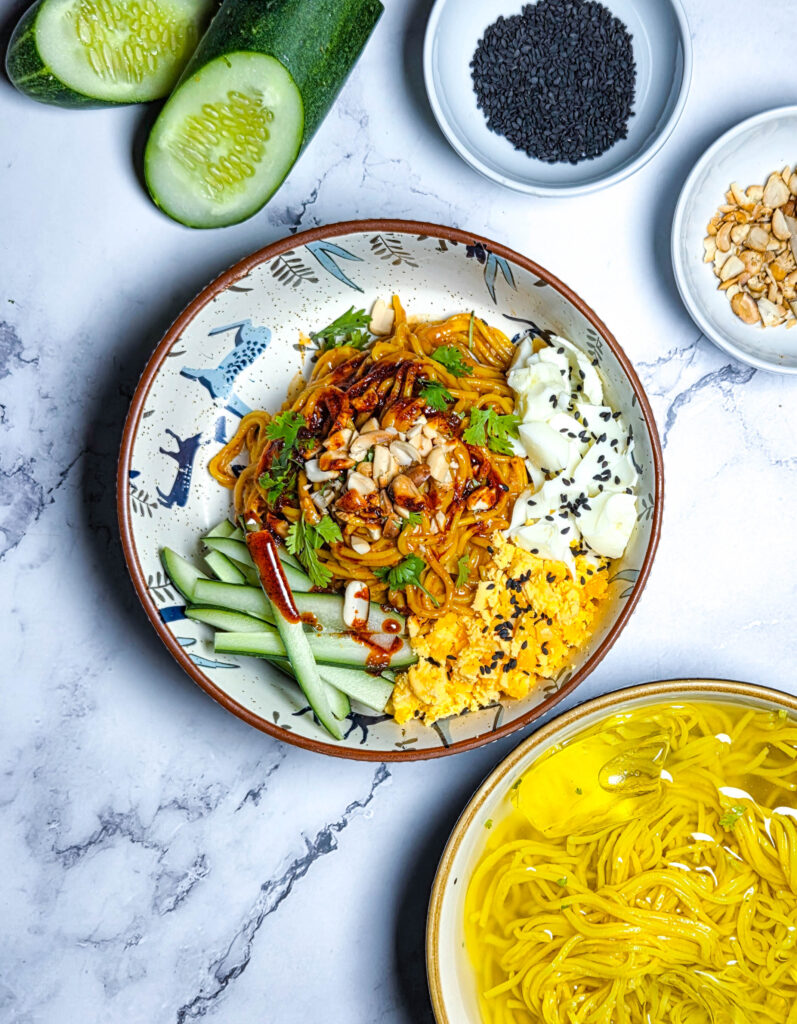 Cold Sesame Noodles in a bowl with peanut sesame chilli oil sauce, garnished with cucumbers, peanuts, coriander, sesame seeds and hardboiled eggs