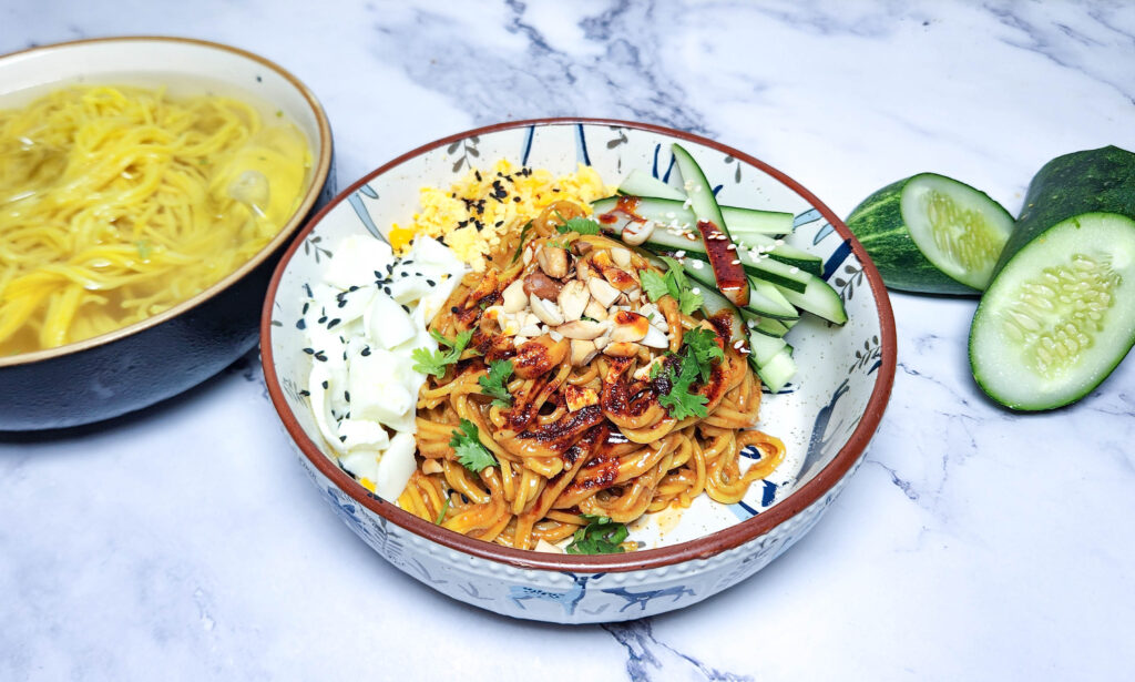 Cold Sesame Noodles in a bowl with peanut sesame chilli oil sauce, garnished with cucumbers, peanuts, coriander, sesame seeds and hardboiled eggs